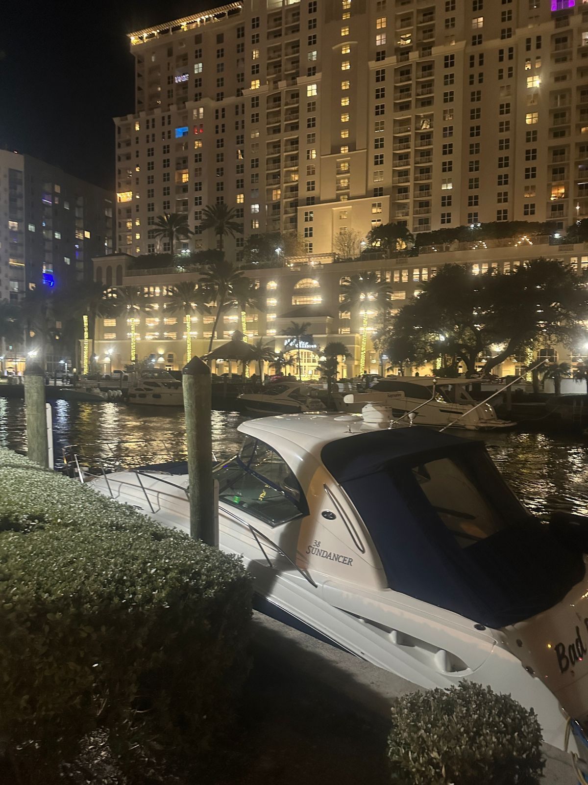 Uma marina à noite com barcos atracados, um edifício iluminado grande ao fundo e um corpo de água.