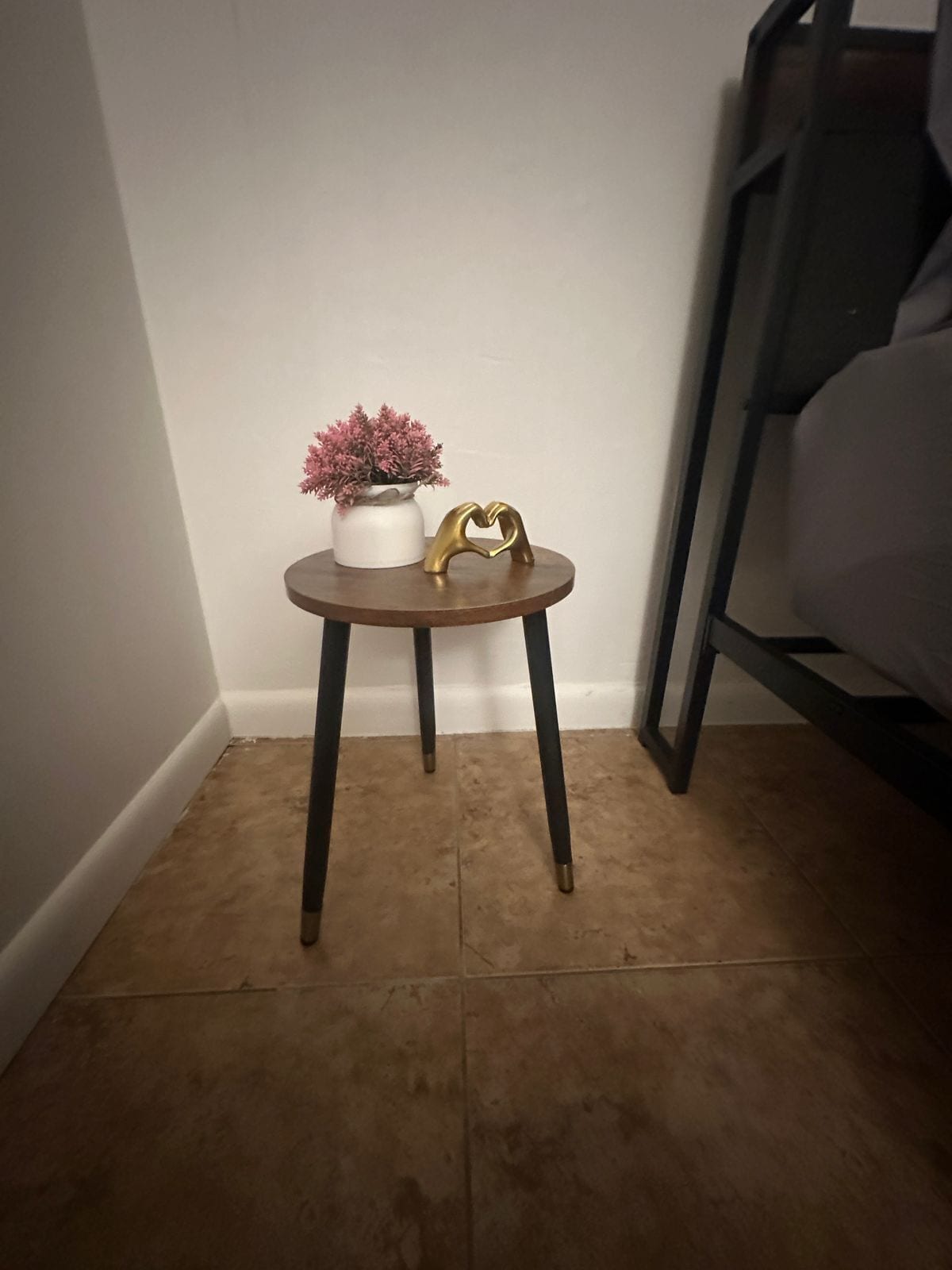 Una pequeña mesa redonda con un jarrón blanco y flores rosadas, un objeto dorado con forma de corazón y un suelo de madera oscura.
