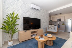 A modern living room with a flat-screen TV, a coffee table, a potted plant, and a kitchen in the background. - Chic Bayahibe 1BR | Portofino | Pool + BBQ Area (Bayahibe)