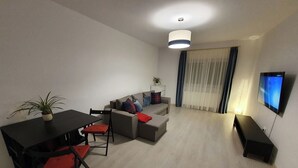 A modern living room with a sofa, a dining table with chairs, a TV mounted on the wall, and a ceiling light. - Sunny and Cozy Apartment (Brașov)