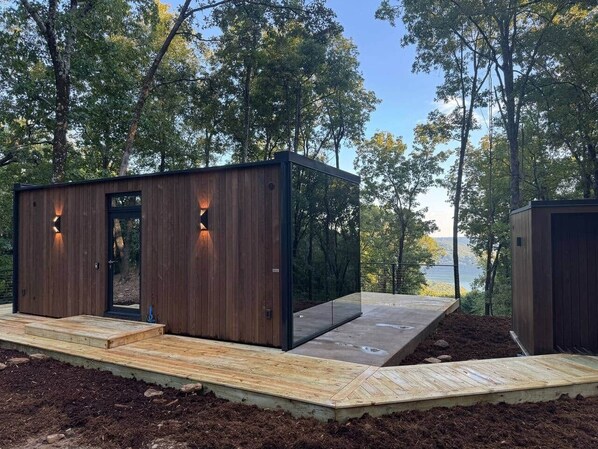 A modern wooden cabin with a deck, built into a forested area with a lake view.
