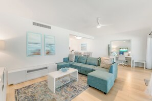 A modern living room with a sectional sofa, a glass coffee table, a white side table, and a desk with a computer. - Casa Caribe #15 - 3 Bed, 3 Bath - Oceanfront, Second Floor (Seven Mile Beach)