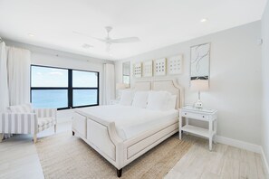 A modern bedroom with a large bed, a side table, a chair, and a view of the sea. - Casa Caribe #23 - 3 Bed, 2 Bath - Oceanfront, Third Floor (Seven Mile Beach)