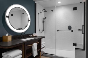 A modern bathroom with a round mirror, dark wooden vanity, white towels, and a glass-enclosed shower area. - Hotel West & Main Conshohocken Philadelphia, Tapestry by Hilton (Conshohocken)