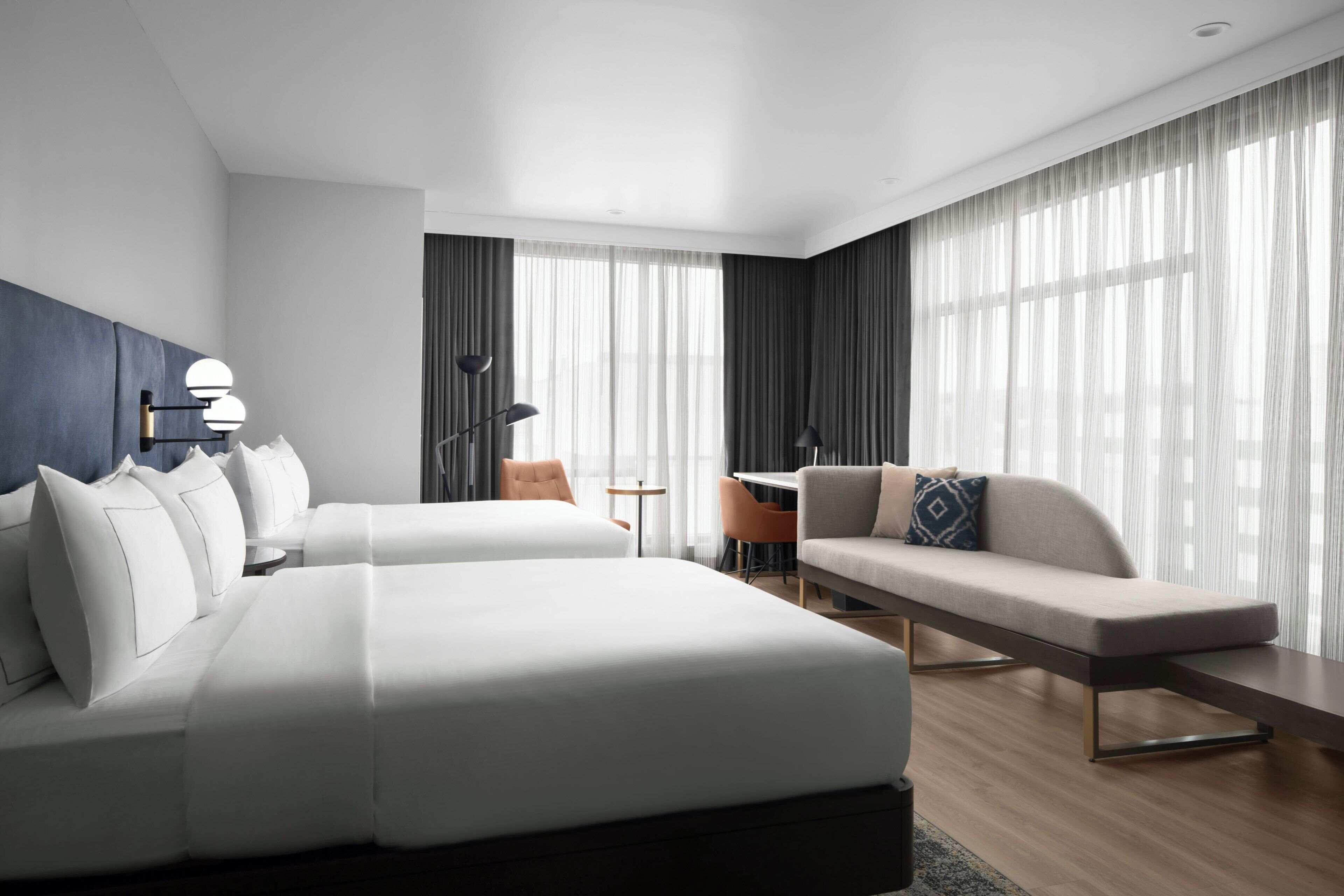 Quarto de hotel moderno com duas camas, um sofá, uma escrivaninha e janelas amplas com cortinas translúcidas.