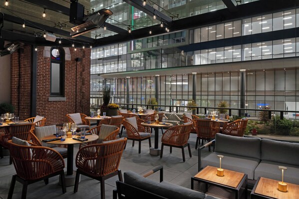 A modern indoor dining area with wicker furniture, a brick accent wall, and large windows. - Hotel West & Main Conshohocken Philadelphia, Tapestry by Hilton (Conshohocken)