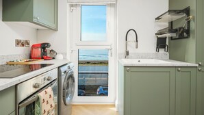 Cocina compacta con lavadora, cafetera y vista al mar a través de una puerta.