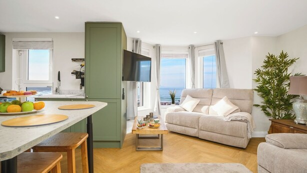 Una sala de estar moderna con un sofá, un comedor con mesa y sillas, una cocina con televisor y vista al mar.