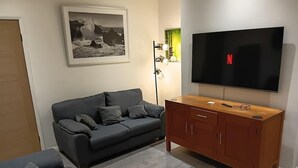 A living room with a grey sofa, a wooden cabinet, a flat-screen TV, a framed picture on the wall, and a floor lamp. - Dale combe,  3 Bedroom House 5 star  on air bnb (Middleton)