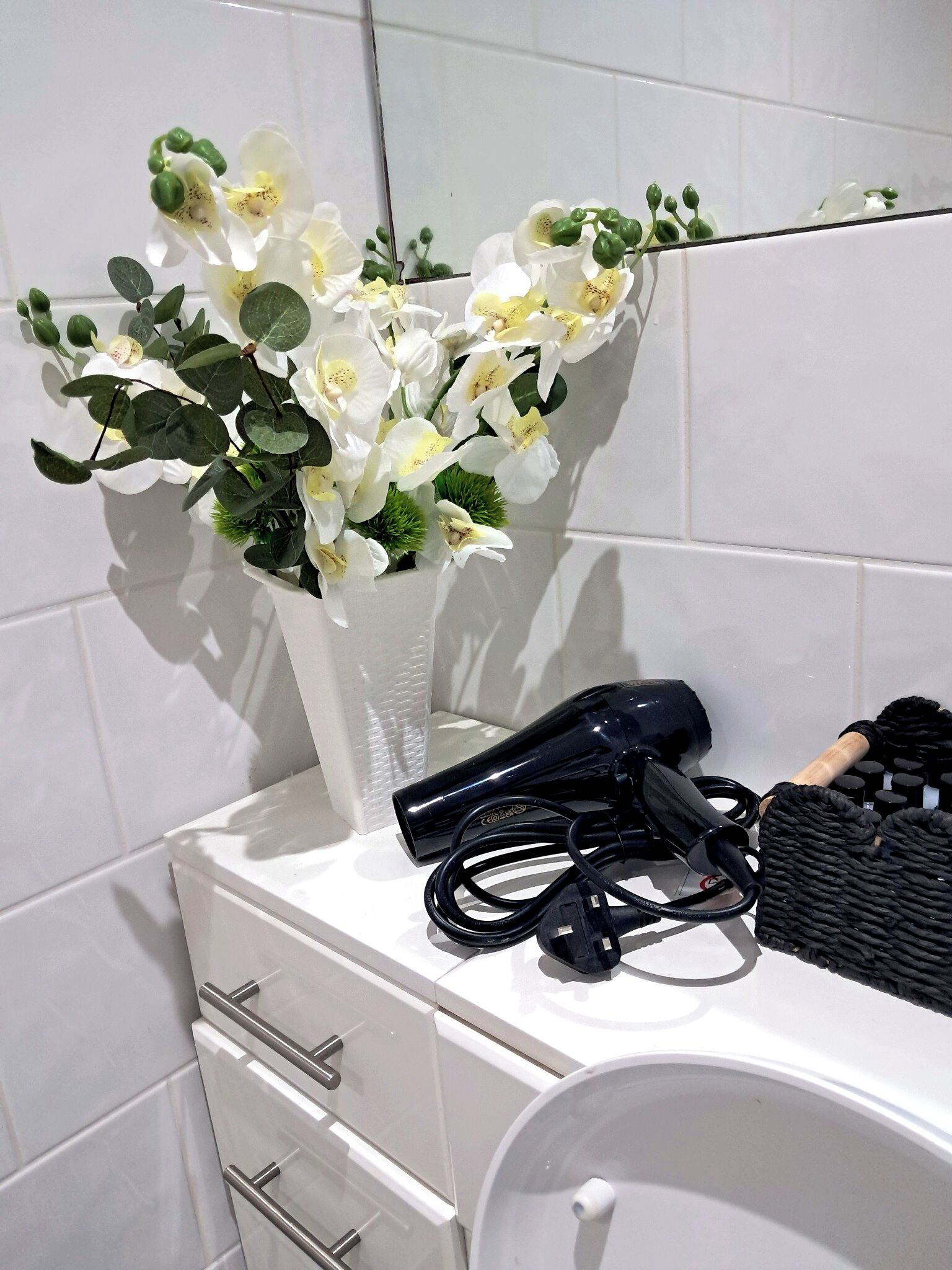 Ett badrum med vita kakelplattor, en vit vas med blommor, en hårtork och en svart handduk.