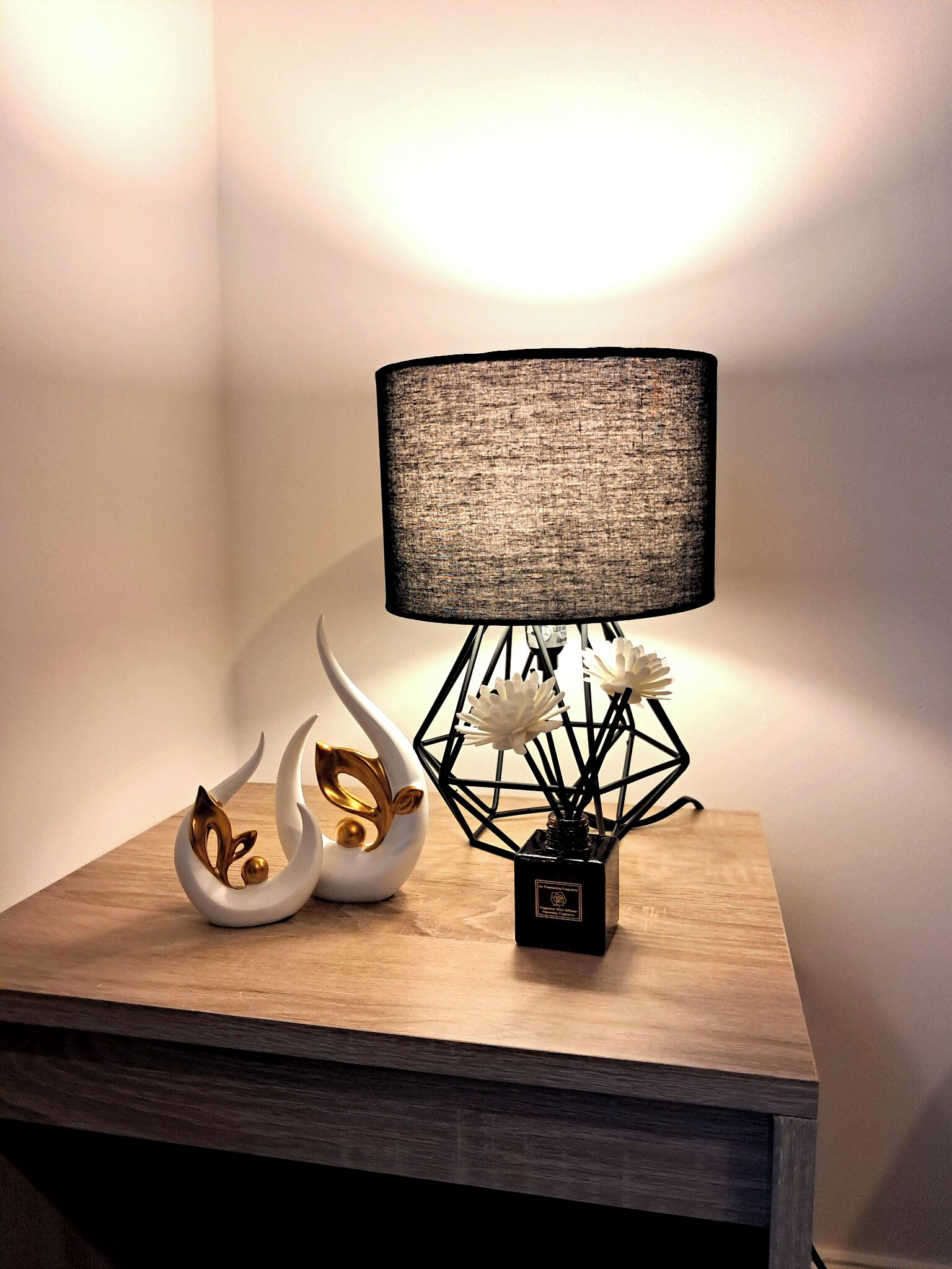 Eine Lampe mit strukturierter Schirm, eine geometrische Vase mit Blumen und eine schwanförmige Dekoration auf einer Holzbodenfläche.