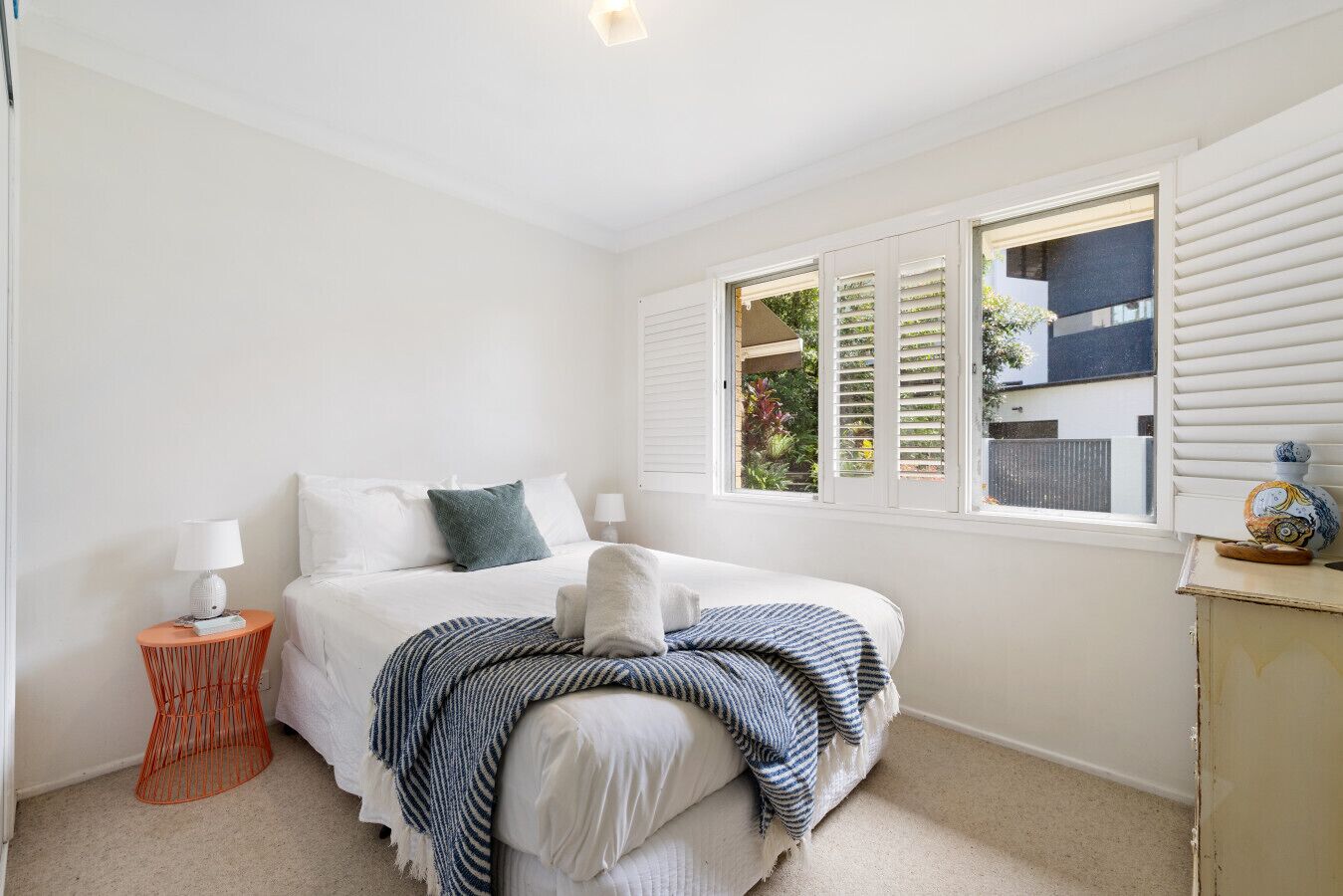 Une chambre à coucher avec un lit, deux tables de chevet, une fenêtre avec des volets et une vue sur la verdure.
