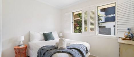 Une chambre à coucher avec un lit, deux tables de chevet, une fenêtre avec des volets et une vue sur la verdure.