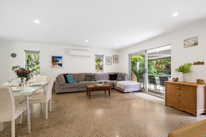 A spacious living area with a sofa, coffee table, and dining table set up. There is a sliding glass door leading to a balcony with greenery outside. - Luci`s Beach Retreat (Coolum Beach)