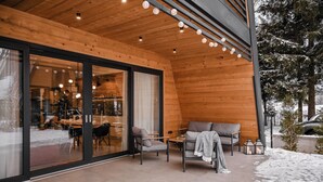 A wooden exterior with large glass doors, a seating area with chairs and a sofa, and a snow-covered ground. - Family mountain retreat with jacuzzi, sauna, playground, and stunning forest views. (Sunger)