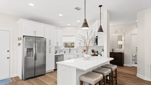 A modern kitchen with white cabinetry, a stainless steel refrigerator, a marble island with bar stools, pendant lighting, and a view to the dining area. - Salt and Sway (Miramar)