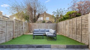 A fenced backyard with a grey sectional sofa on artificial grass, stone paving, and a brick house in the background. - The Luxury Secret Garden Retreat 3 bed 2 Bath (London)