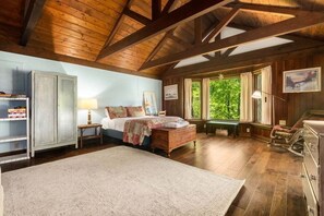 Ein rustikales Schlafzimmer mit Holzbalkendecke, einem Bett mit Quilt, einem Nachttisch, einem Schaukelstuhl und einem Fenster mit Blick auf Bäume.
