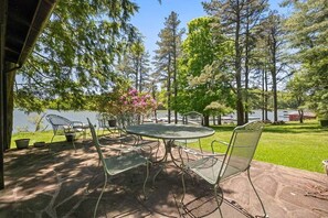 Eine Terrasse im Freien mit einem runden Tisch und Metallstühlen, umgeben von Bäumen und Blick auf einen See.