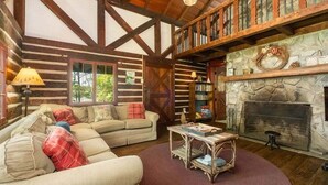 Ein Interieur einer Blockhütte mit Kamin aus Naturstein, Holzbalken, einer Couch und einem Bücherregal.