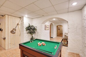 A pool table with balls, a guitar, and a plant in a room with white walls and tiled floors.