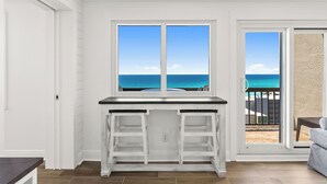 Une pièce avec un bureau blanc, une vue sur l’océan par la fenêtre et une porte-fenêtre coulissante donnant sur un balcon.