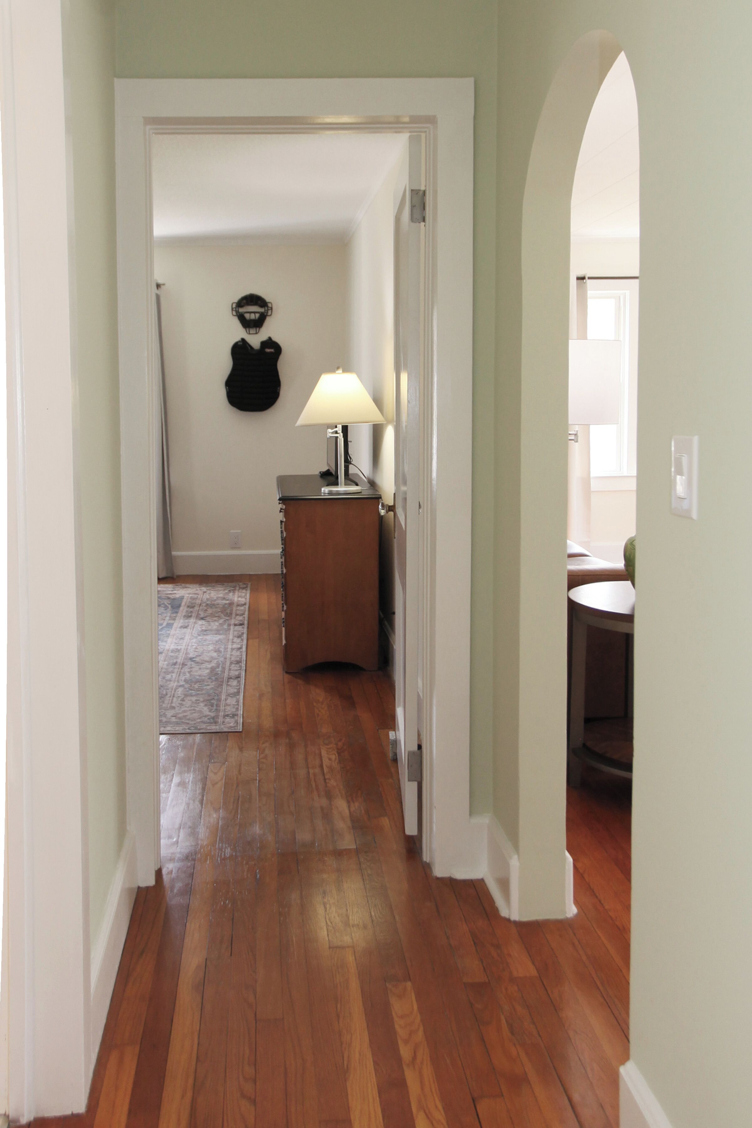 A hallway with wooden flooring, a door leading to a room with a lamp, and a coat rack.