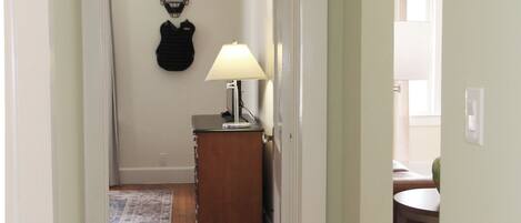 A hallway with wooden flooring, a door leading to a room with a lamp, and a coat rack.