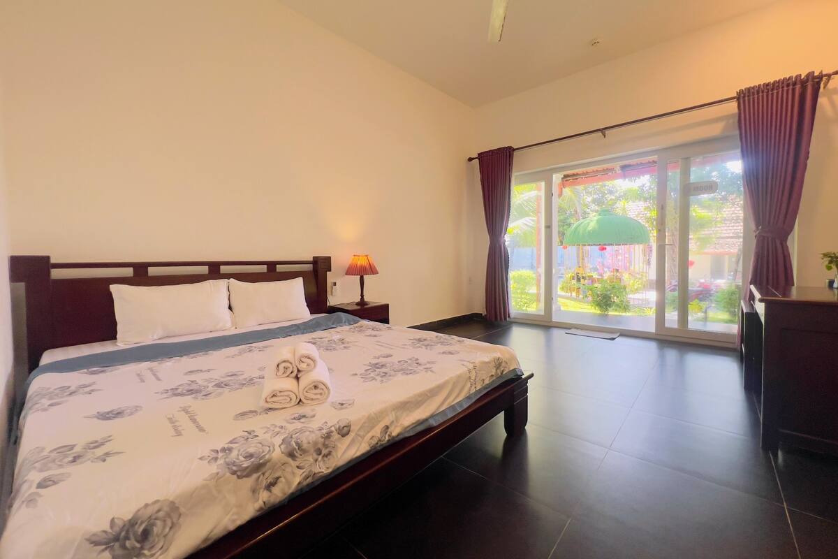 A hotel room with a bed, a nightstand, a lamp, a window with curtains, and a view of the outside.