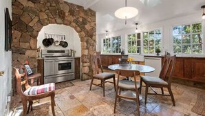 Un arc en pierre mène à une cuisine équipée d’une cuisinière, d’un four et de casseroles suspendues. On y trouve également un coin repas avec une table ronde et des chaises, ainsi qu’une fenêtre donnant sur un paysage verdoyant.