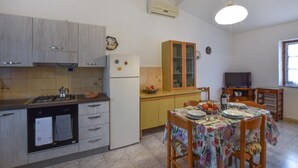 Une cuisine équipée d’une cuisinière, d’un réfrigérateur et de placards. On y trouve une table à manger avec des chaises, et un téléviseur est visible au fond.