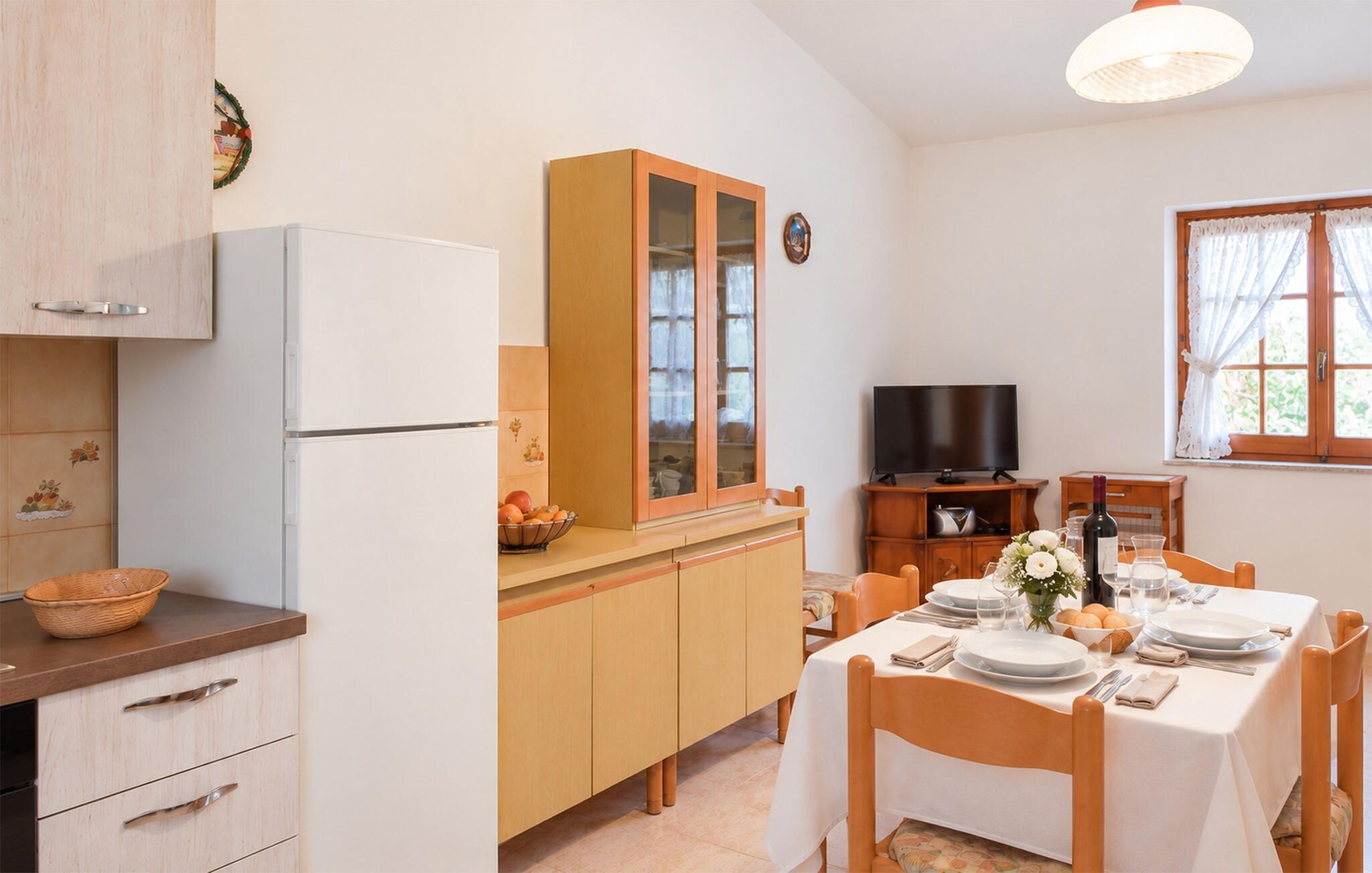 Eine kleine Küche mit Kühlschrank, Holzschränken, einem Esstisch für zwei Personen und einem Fernseher.