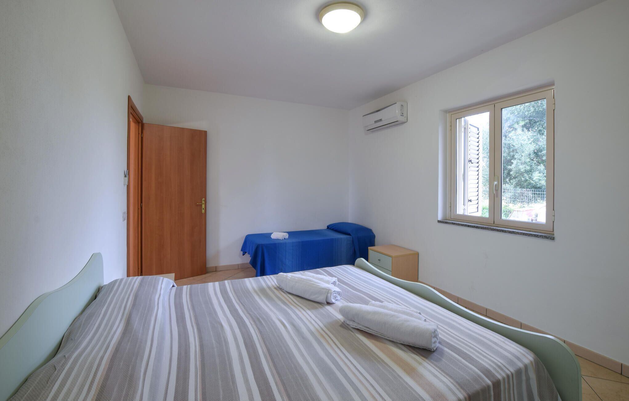 A bedroom with a bed, a small table, a chair, a window, and an air conditioning unit.