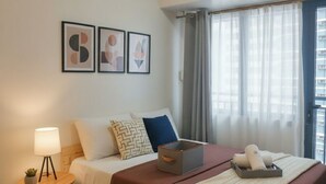 A hotel room with a bed, a bedside table with a lamp, a small plant, a decorative box, and a window with curtains. - Makati City Central Stay Apartments (Makati)