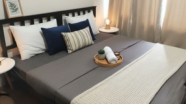 A neatly made bed with a dark-colored mattress, a white and blue pillow arrangement, a small plant, and a rolled towel on a tray. - Makati City Central Stay Apartments (Makati)