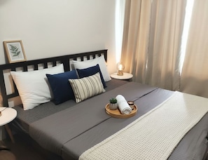 A neatly made bed with a dark-colored mattress, a white and blue pillow arrangement, a small plant, and a rolled towel on a tray. - Makati City Central Stay Apartments (Makati)