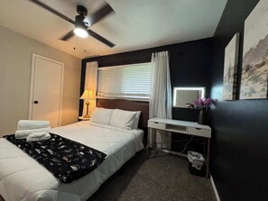 Ein Hotelzimmer mit Bett, Schreibtisch, Deckenventilator, Lampe und einem fenster mit Vorhängen.