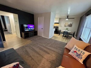 Ein Wohnzimmer mit Flachbildfernseher, einer Couch mit einem Texas-Dekokissen, einem Essbereich mit Tisch und Stühlen sowie einer Küche mit Herd und Schränken.