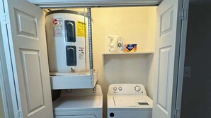Un lavadero con lavadora y secadora apiladas, un estante con cafetera y detergente, y una puerta que da a otra habitación.