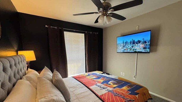 Una habitación de hotel con cama, ventilador de techo, televisor en la pared y ventana con cortinas.