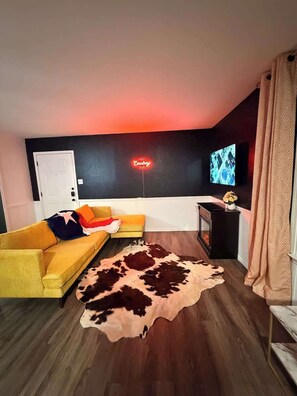 Olohuone, jossa on keltainen sohva, lehmännahkamatto, televisio ja neonvalomainos.
