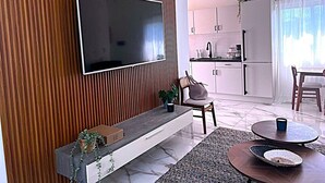 A modern living room with a flat-screen TV, wooden panel wall, a bench with plants, a dining area with a table and chairs, and a kitchen in the background. - Surf View Penthouse @ Encuentro (Cabarete)