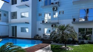 A modern building with a swimming pool, outdoor seating, and a small palm tree. - Surf View Penthouse @ Encuentro (Cabarete)
