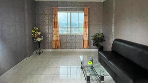 A modern living room with a black sofa, a glass coffee table, a potted plant, a flower arrangement, and a view through a window with curtains.
