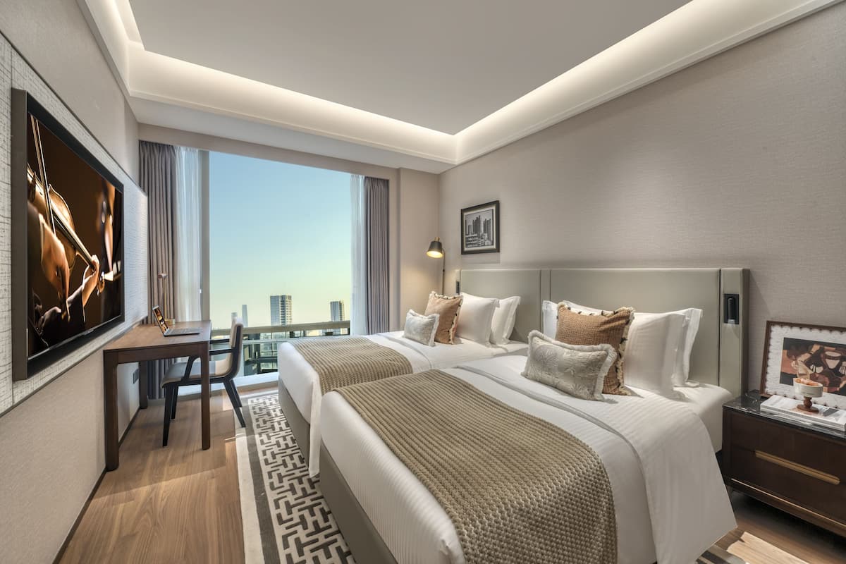 A hotel room with two beds, a desk, a TV, and a view of the city.