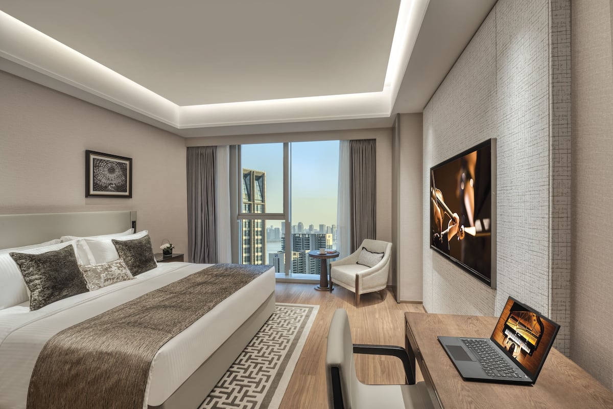 A modern hotel room with a large bed, a desk with a laptop, a TV, and a city view.