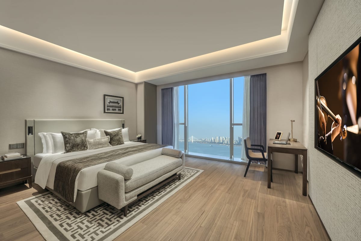 A modern hotel room with a large bed, a desk, a TV, and a view of a cityscape.
