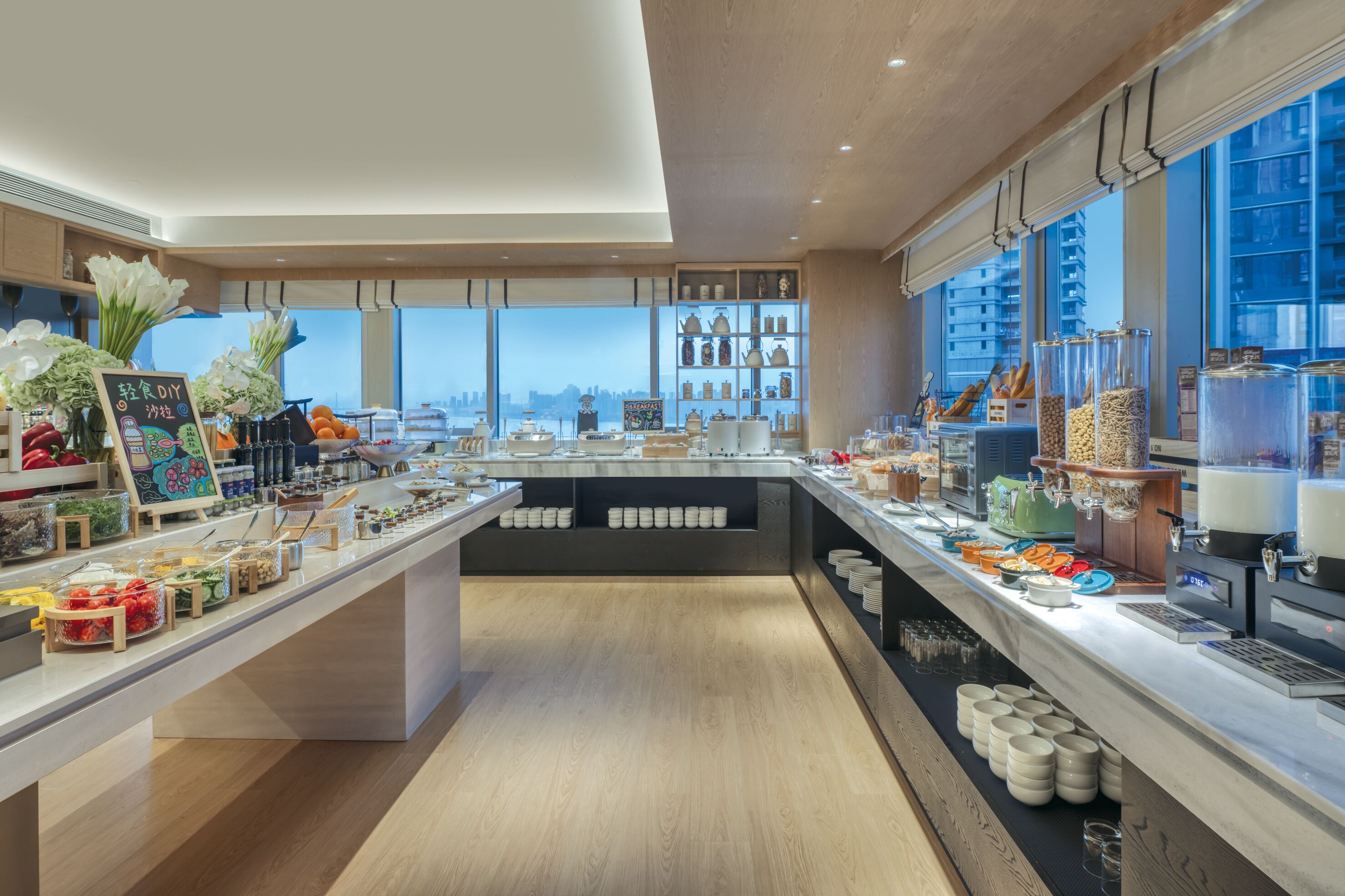 A modern cafe with a long counter, various food items, and a city view.