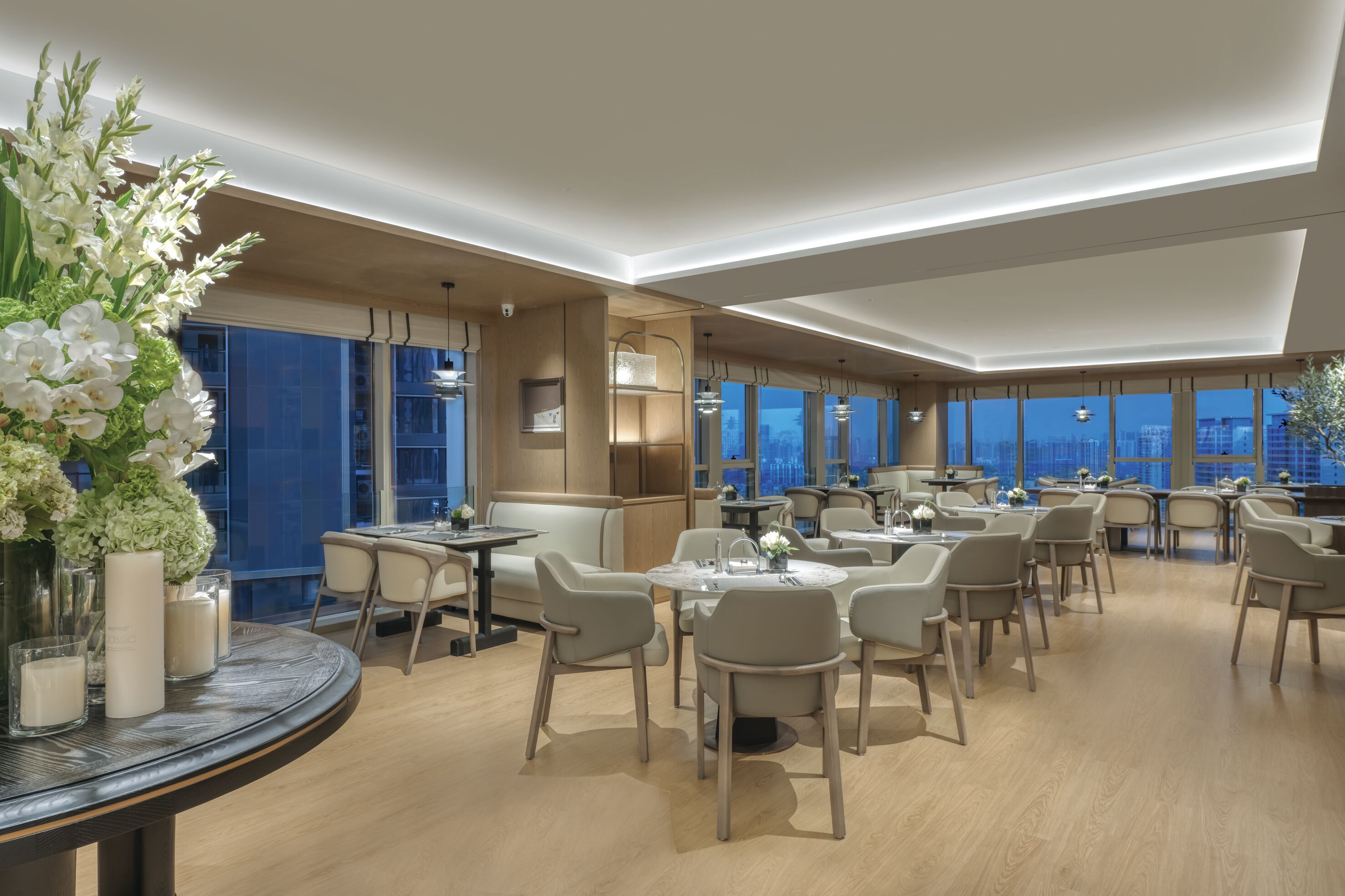 A modern hotel dining area with round tables and chairs, large windows, and a view of the cityscape.