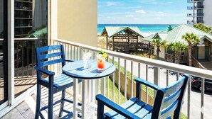 Un balcon avec deux chaises bleues et une petite table, donnant sur une plage avec une structure en bois et des palmiers.